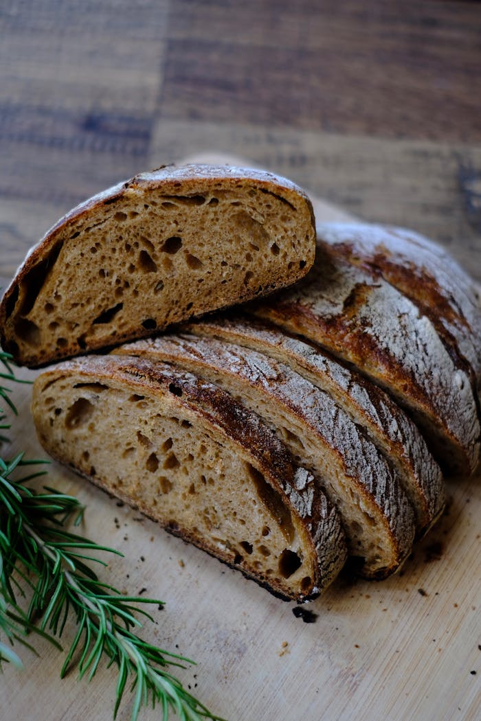 Home Close-up of a sliced, crusty sourdough loaf with fresh rosemary on a wooden surface.